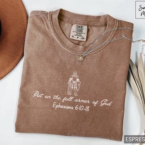 Puede incluir: Una camiseta marr&oacute;n con un gr&aacute;fico blanco de un caballero con armadura y el texto "Ponte la armadura completa de Dios Efesios 6:10-18".