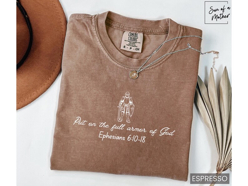 Puede incluir: Una camiseta marr&oacute;n con un gr&aacute;fico blanco de un caballero con armadura y el texto "Ponte la armadura completa de Dios Efesios 6:10-18".