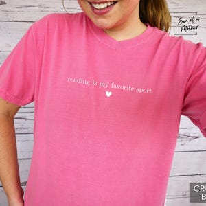 Puede incluir: Una camiseta rosa con el texto "reading is my favorite sport" y un peque&ntilde;o coraz&oacute;n blanco. La camiseta es de color liso y tiene mangas cortas. La esquina inferior derecha de la camiseta tiene el texto "CRUNCH-BERRY".