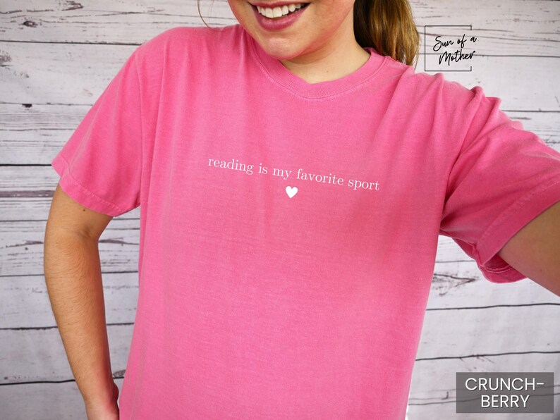 Puede incluir: Una camiseta rosa con el texto "reading is my favorite sport" y un peque&ntilde;o coraz&oacute;n blanco. La camiseta es de color liso y tiene mangas cortas. La esquina inferior derecha de la camiseta tiene el texto "CRUNCH-BERRY".
