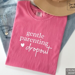 Puede incluir: Una camiseta rosa con las palabras "gentle parenting dropout" en escritura blanca y un peque&ntilde;o coraz&oacute;n blanco. La camiseta est&aacute; doblada sobre unos vaqueros azules, con un sombrero beige y un collar dorado visibles.