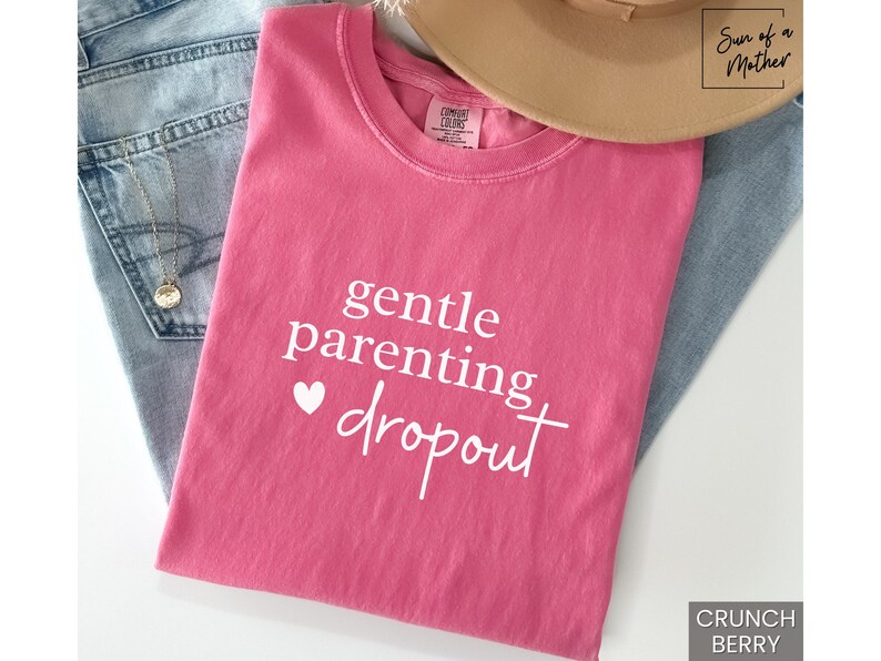 Puede incluir: Una camiseta rosa con las palabras "gentle parenting dropout" en escritura blanca y un peque&ntilde;o coraz&oacute;n blanco. La camiseta est&aacute; doblada sobre unos vaqueros azules, con un sombrero beige y un collar dorado visibles.