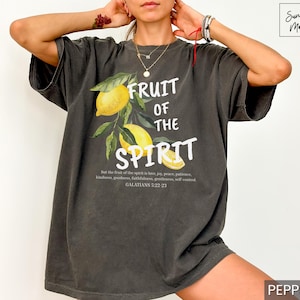 Op de afbeelding: Een donkergrijs T-shirt met een afbeelding van citroenen en de tekst "Fruit of the Spirit" en een bijbelvers uit Galaten 5:22-21.