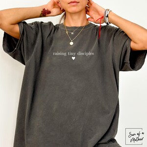 Op de afbeelding: Donkergrijs t-shirt met de tekst "raising tiny disciples" en een kleine hartafbeelding. Het shirt heeft een vintage, gewassen uitstraling. De merknaam "Sun of a Mother" staat rechtsonder.