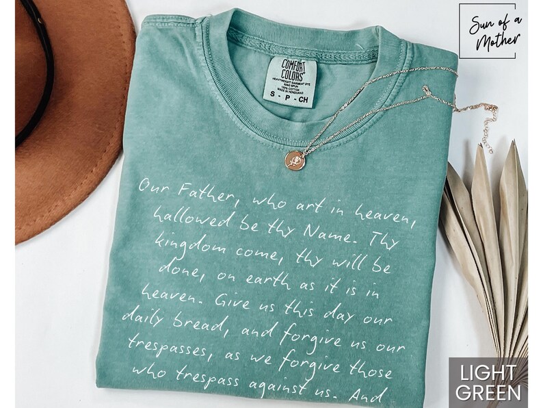 May include: Light green t-shirt with the Lord's Prayer written in white script. The shirt has a distressed look and is made of a soft, comfortable fabric. The text on the shirt reads: "Our Father, who art in heaven, hallowed be thy Name. Thy kingdom come, thy will be done, on earth as it is in heaven. Give us this day our daily bread, and forgive us our trespasses, as we forgive those who trespass against us. And"