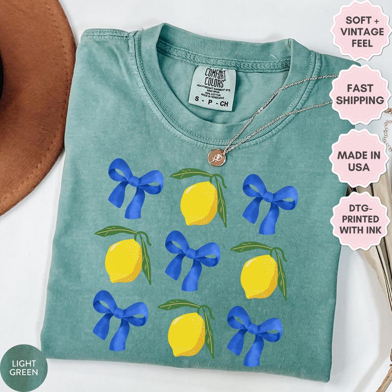 K&ouml;nnte beinhalten: Hellgr&uuml;nes T-Shirt mit einem Muster aus Zitronen und blauen Schleifen. Das Shirt hat einen Used-Look und ist in den USA hergestellt. Der Text "Comfort Colors" ist auf dem Shirt sichtbar.