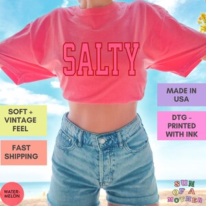 May include: A pink cropped t-shirt with the word "SALTY" printed in red on the front. The shirt is made in the USA and printed with ink. The model is wearing the shirt with denim shorts.