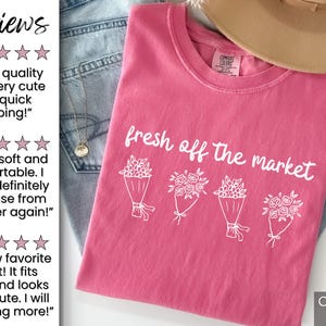 May include: A pink t-shirt with the phrase "fresh off the market" and illustrations of flower bouquets. The shirt is folded on top of blue jeans. A tan hat is visible in the background.