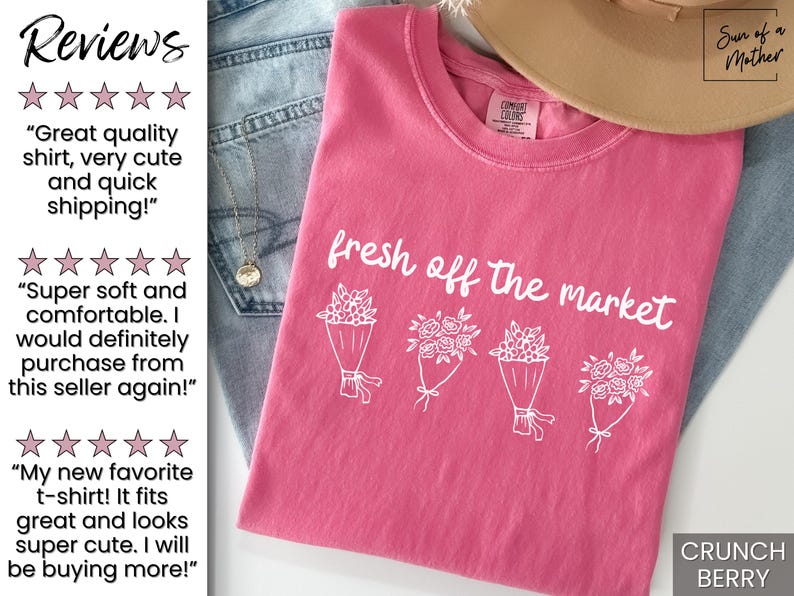 May include: A pink t-shirt with the phrase "fresh off the market" and illustrations of flower bouquets. The shirt is folded on top of blue jeans. A tan hat is visible in the background.