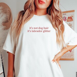 May include: White t-shirt with the text "it's not dog hair it's labrador glitter" in red. The shirt is a casual style with short sleeves and a crew neck. The text is centered on the front of the shirt.