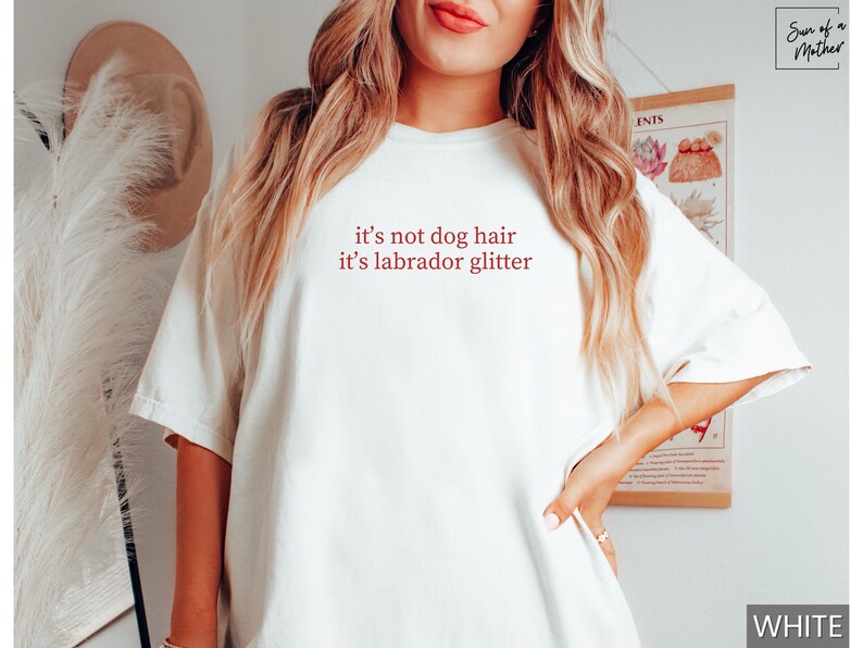 May include: White t-shirt with the text "it's not dog hair it's labrador glitter" in red. The shirt is a casual style with short sleeves and a crew neck. The text is centered on the front of the shirt.