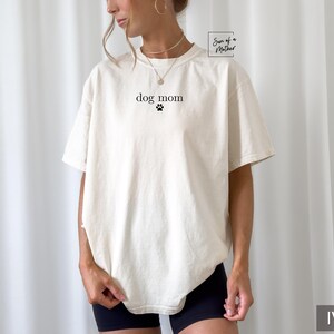 May include: A white t-shirt with the text "dog mom" and a paw print in black. The shirt is being worn by a person with a light brown complexion. The background is a white wall.