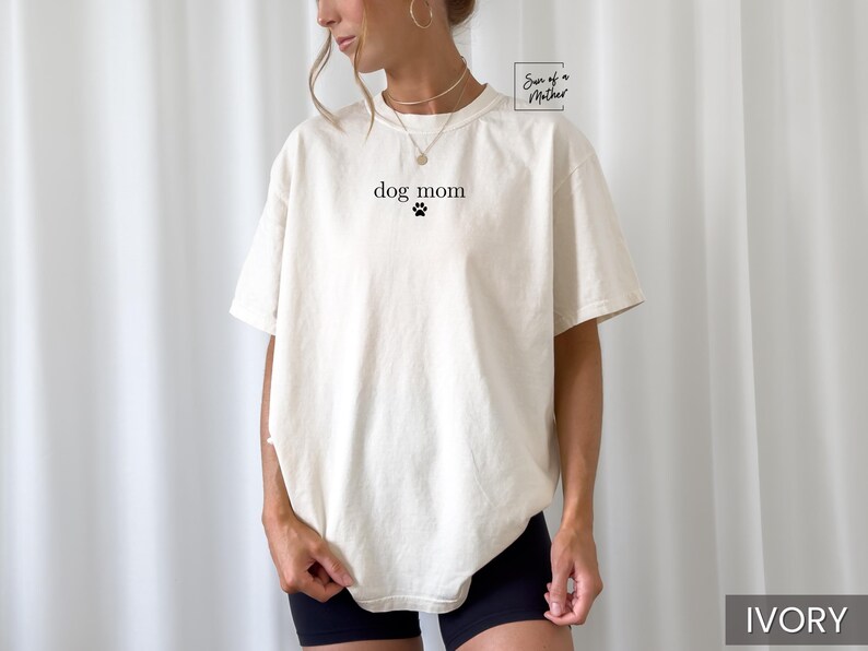 May include: A white t-shirt with the text "dog mom" and a paw print in black. The shirt is being worn by a person with a light brown complexion. The background is a white wall.