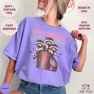 Op de afbeelding: Paars T-shirt met een afbeelding van drie wasberen in een prullenbak. De tekst "Stay Trashy" staat boven de wasberen. Het shirt is gemaakt in de VS en bedrukt met DTG-inkt.