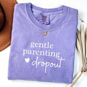 Puede incluir: Camiseta lila con la frase "gentle parenting dropout" en blanco. Un collar dorado con un colgante descansa sobre la camiseta. Un sombrero marr&oacute;n y hojas de palma secas en el fondo.