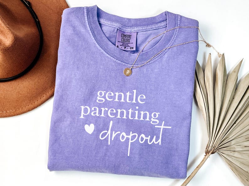 Puede incluir: Camiseta lila con la frase "gentle parenting dropout" en blanco. Un collar dorado con un colgante descansa sobre la camiseta. Un sombrero marr&oacute;n y hojas de palma secas en el fondo.