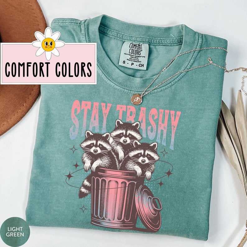 Op de afbeelding: Lichtgroen T-shirt met een afbeelding van drie wasberen in een prullenbak. De tekst "Stay Trashy" staat boven de wasberen. Het shirt is van het merk "Comfort Colors".