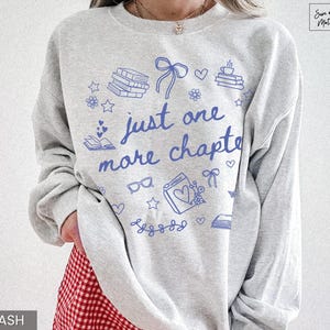 May include: Light gray sweatshirt with blue illustrations of books, hearts, and bows. The text "just one more chapter" is written in blue script. The sweatshirt is a casual style, perfect for book lovers.