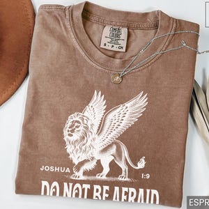 May include: Espresso-colored t-shirt featuring a winged lion graphic with the text "Joshua 1:9 DO NOT BE AFRAID." A silver necklace with a pendant rests on the shirt. The shirt is a Comfort Colors brand, size S-CH.