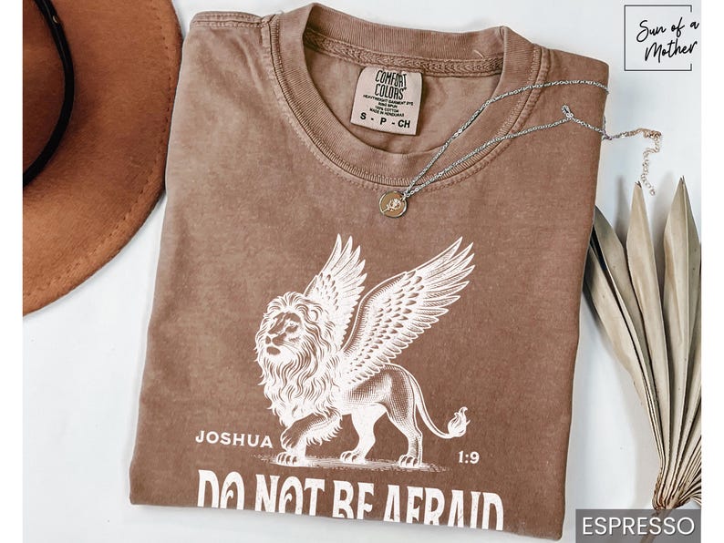 May include: Espresso-colored t-shirt featuring a winged lion graphic with the text "Joshua 1:9 DO NOT BE AFRAID." A silver necklace with a pendant rests on the shirt. The shirt is a Comfort Colors brand, size S-CH.