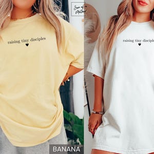 Op de afbeelding: Twee T-shirts, &eacute;&eacute;n in bananengeel en &eacute;&eacute;n in wit, elk met de tekst "raising tiny disciples" boven een zwart hart. De shirts hebben korte mouwen en lijken van een zacht materiaal te zijn.