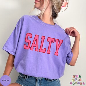 May include: A purple t-shirt with the word "SALTY" printed in pink letters with a red outline. The shirt is being worn by a person.