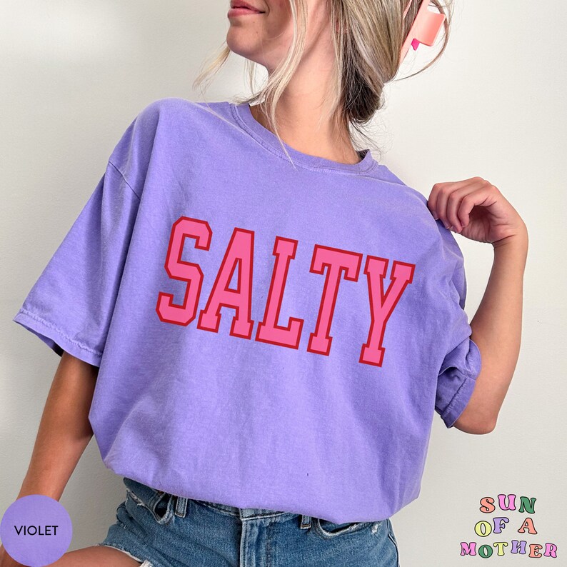 May include: A purple t-shirt with the word "SALTY" printed in pink letters with a red outline. The shirt is being worn by a person.