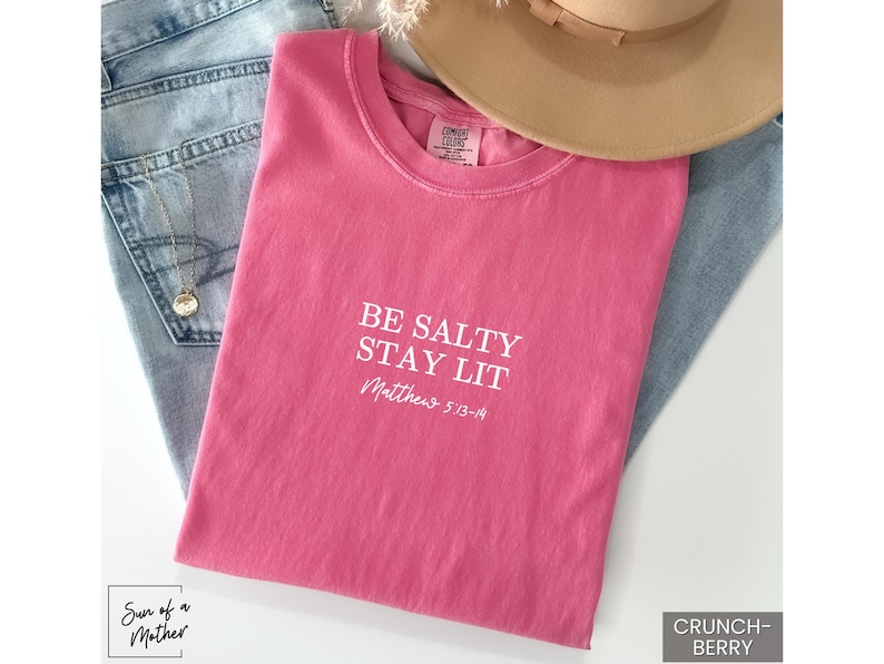 Puede incluir: Camiseta rosa con el texto "Be Salty Stay Lit" y "Matthew 5:13-14" en letras blancas.