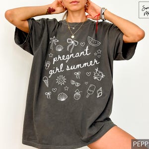 May include: Dark gray t-shirt with the phrase "pregnant girl summer" in white script, surrounded by summer-themed doodles. The shirt has a relaxed fit and short sleeves. The word "pepper" is in the bottom right corner.