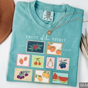 May include: A teal blue t-shirt with the text "FRUIT of the SPIRIT" and a graphic of nine colorful fruit stamps. The stamps include strawberries, cherries, a pineapple, blueberries, a watermelon, and oranges. The text "GAL 42" and "5:22-23" are also included on the shirt.