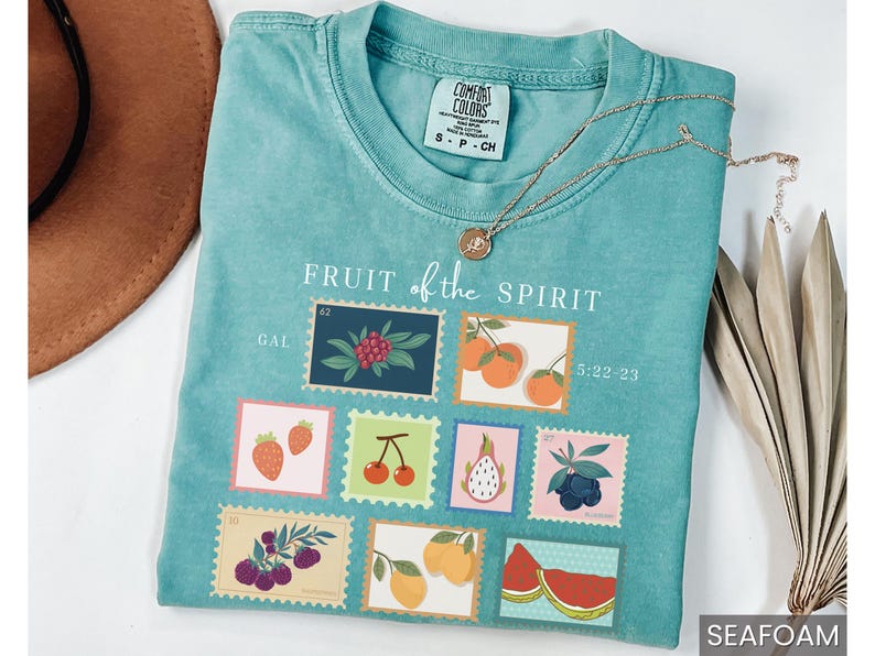 May include: A teal blue t-shirt with the text "FRUIT of the SPIRIT" and a graphic of nine colorful fruit stamps. The stamps include strawberries, cherries, a pineapple, blueberries, a watermelon, and oranges. The text "GAL 42" and "5:22-23" are also included on the shirt.