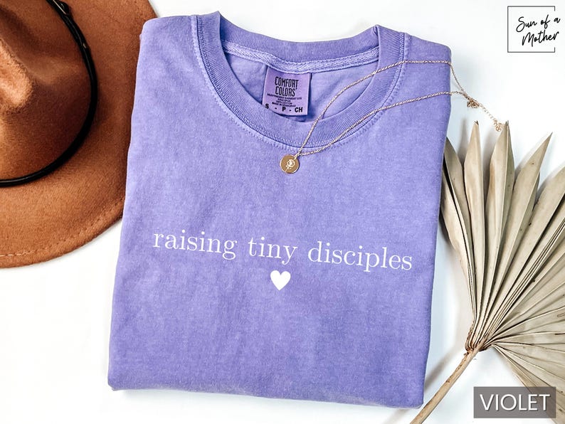 Op de afbeelding: Een violet Comfort Colors T-shirt met de tekst "raising tiny disciples" en een hartje. Een gouden ketting en een bruine hoed zijn ook op de afbeelding te zien. Het shirt is maat S.