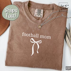 May include: Espresso-colored t-shirt with the words "football mom" in white script, along with a white bow graphic. The shirt has a crew neck and short sleeves. A silver necklace with a pendant is draped across the shirt.