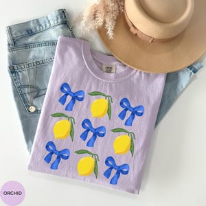 K&ouml;nnte beinhalten: Ein lavendelfarbenes T-Shirt mit einem grafischen Aufdruck von Zitronen und blauen Schleifen. Der Text "COMFORT COLORS" ist auf dem Shirt aufgedruckt.