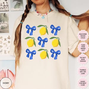 K&ouml;nnte beinhalten: Ein wei&szlig;es T-Shirt mit einem Zitronen- und blauen Schleifendesign. Das Shirt hat Pflegehinweise, die auf der rechten Seite des Bildes aufgedruckt sind. Der Text lautet: "Maschinenw&auml;sche kalt, Kleidungsst&uuml;ck auf links drehen, Design nicht b&uuml;geln, Trocknergeeignet bei niedriger Temperatur, zum Trocknen aufh&auml;ngen."