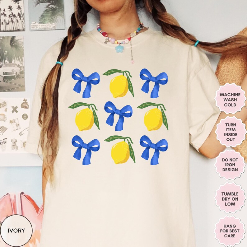 K&ouml;nnte beinhalten: Ein wei&szlig;es T-Shirt mit einem Zitronen- und blauen Schleifendesign. Das Shirt hat Pflegehinweise, die auf der rechten Seite des Bildes aufgedruckt sind. Der Text lautet: "Maschinenw&auml;sche kalt, Kleidungsst&uuml;ck auf links drehen, Design nicht b&uuml;geln, Trocknergeeignet bei niedriger Temperatur, zum Trocknen aufh&auml;ngen."