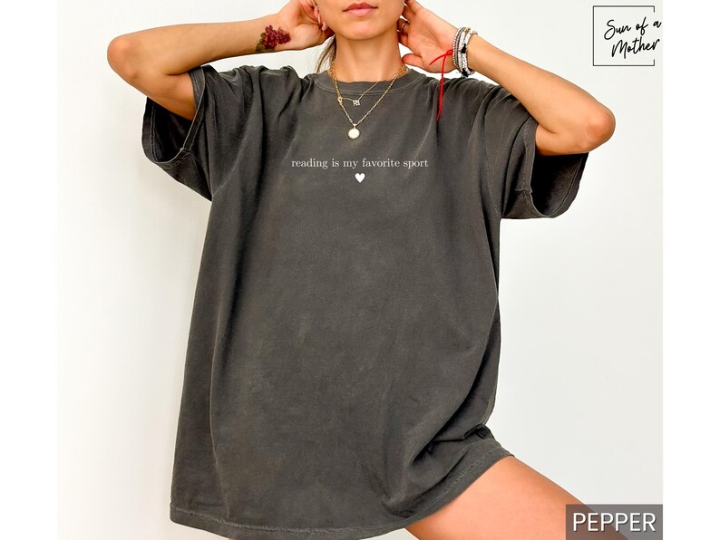 Puede incluir: Camiseta gris oscuro de gran tama&ntilde;o con el texto "reading is my favorite sport" y un peque&ntilde;o coraz&oacute;n. La camiseta tiene mangas cortas y un corte holgado, perfecta para uso casual.