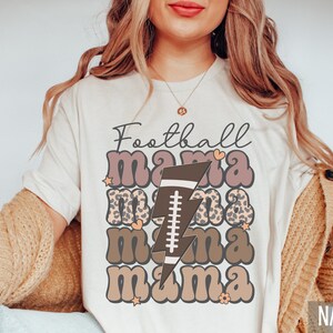 Puede incluir: Camiseta blanca con el texto "Football mama" en letras marrones y rosas con un rayo marr&oacute;n y un gr&aacute;fico de f&uacute;tbol. La camiseta es de color natural.