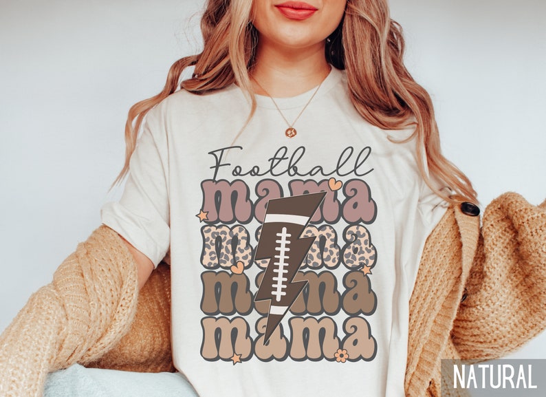 Puede incluir: Camiseta blanca con el texto "Football mama" en letras marrones y rosas con un rayo marr&oacute;n y un gr&aacute;fico de f&uacute;tbol. La camiseta es de color natural.