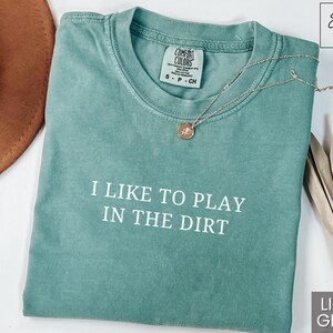 May include: Light green t-shirt with the text "I like to play in the dirt" printed in white. The shirt is made of a soft, comfortable material.