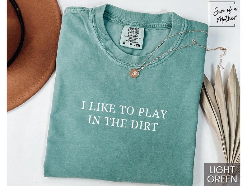 May include: Light green t-shirt with the text "I like to play in the dirt" printed in white. The shirt is made of a soft, comfortable material.
