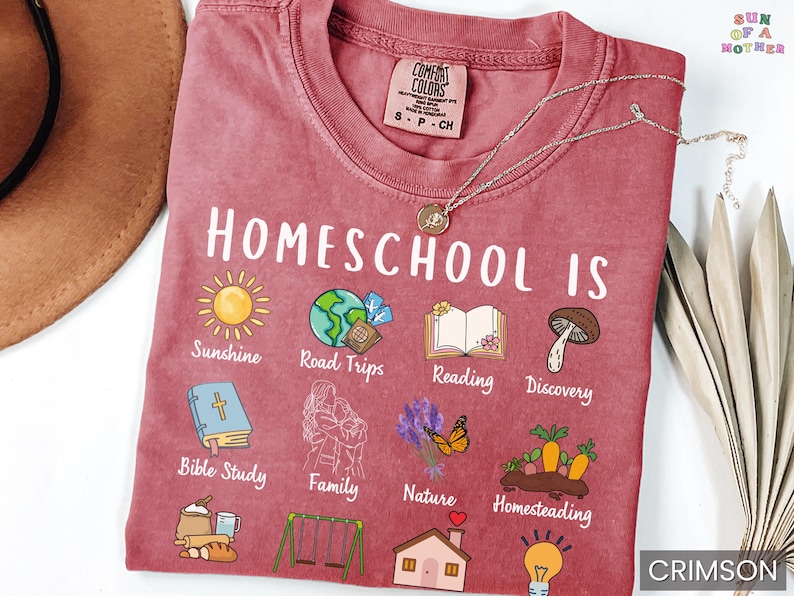 K&ouml;nnte beinhalten: Karmesinrotes T-Shirt mit der Aufschrift "Homeschool Is" und verschiedenen Illustrationen, darunter eine Sonne, ein Globus, ein Buch und ein Pilz. Das Shirt zeigt auch den Text "Sunshine, Road Trips, Reading, Discovery, Bible Study, Family, Nature, Homesteading."