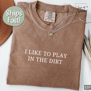 May include: A brown t-shirt with the text "I LIKE TO PLAY IN THE DIRT" printed in white. The shirt is made of a soft, comfortable material and has a distressed look.