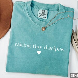 Op de afbeelding: Zeegroen t-shirt met de tekst "raising tiny disciples" en een wit hart. Een gouden ketting met een hanger ligt op het shirt. Een bruine hoed en een gedroogde plant staan op de achtergrond.