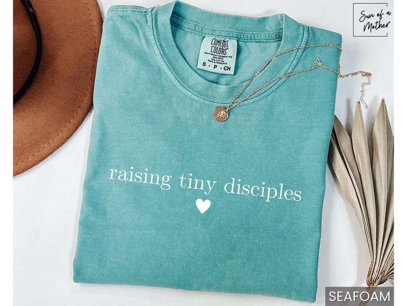 Op de afbeelding: Zeegroen t-shirt met de tekst "raising tiny disciples" en een wit hart. Een gouden ketting met een hanger ligt op het shirt. Een bruine hoed en een gedroogde plant staan op de achtergrond.