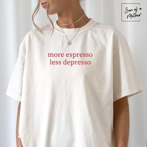 Op de afbeelding: Ivoor kleurig t-shirt met de rode tekst "more espresso less depresso". Het shirt heeft een relaxte pasvorm en korte mouwen. Het merklogo "Sun of a Mother" staat rechtsboven.