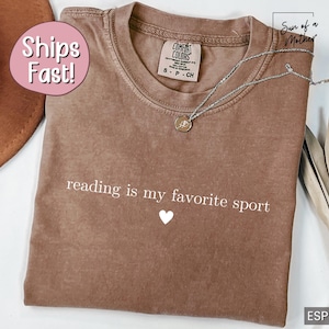 Puede incluir: Camiseta marr&oacute;n con la frase "reading is my favorite sport" y un peque&ntilde;o coraz&oacute;n blanco. La camiseta tiene cuello redondo y mangas cortas. Un collar plateado con un colgante est&aacute; sobre la camiseta.