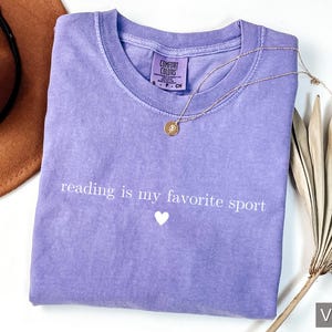 Puede incluir: Una camiseta violeta con la frase "reading is my favorite sport" en blanco y un peque&ntilde;o coraz&oacute;n blanco. Tambi&eacute;n se ven un collar dorado y un sombrero marr&oacute;n.