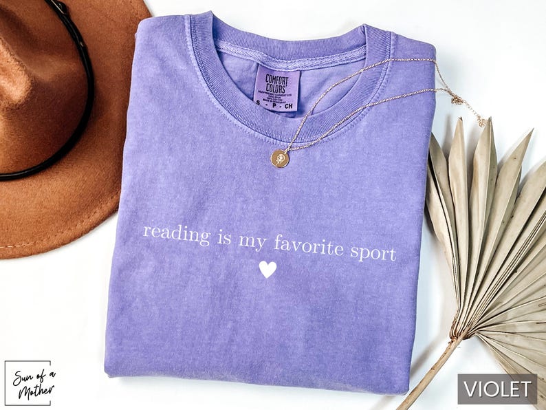 Puede incluir: Una camiseta violeta con la frase "reading is my favorite sport" en blanco y un peque&ntilde;o coraz&oacute;n blanco. Tambi&eacute;n se ven un collar dorado y un sombrero marr&oacute;n.