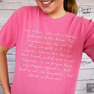 May include: Pink t-shirt with white text that reads "Our Father, who art in heaven, hallowed be thy Name. Thy kingdom come, thy will be done, on earth as it is in heaven. Give us this day our daily bread, and forgive us our trespasses, as we forgive those who trespass against us. And lead us not into temptation, but deliver us from evil."
